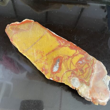 A flat, irregularly shaped piece of yellow and red jasper with some translucent and rough edges, placed on a reflective black surface.