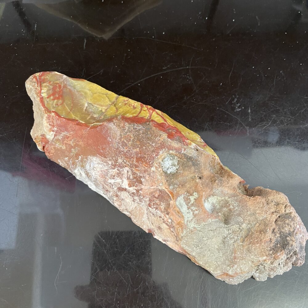 A rough, elongated rock with red, yellow, and white coloration lies on a reflective black surface.