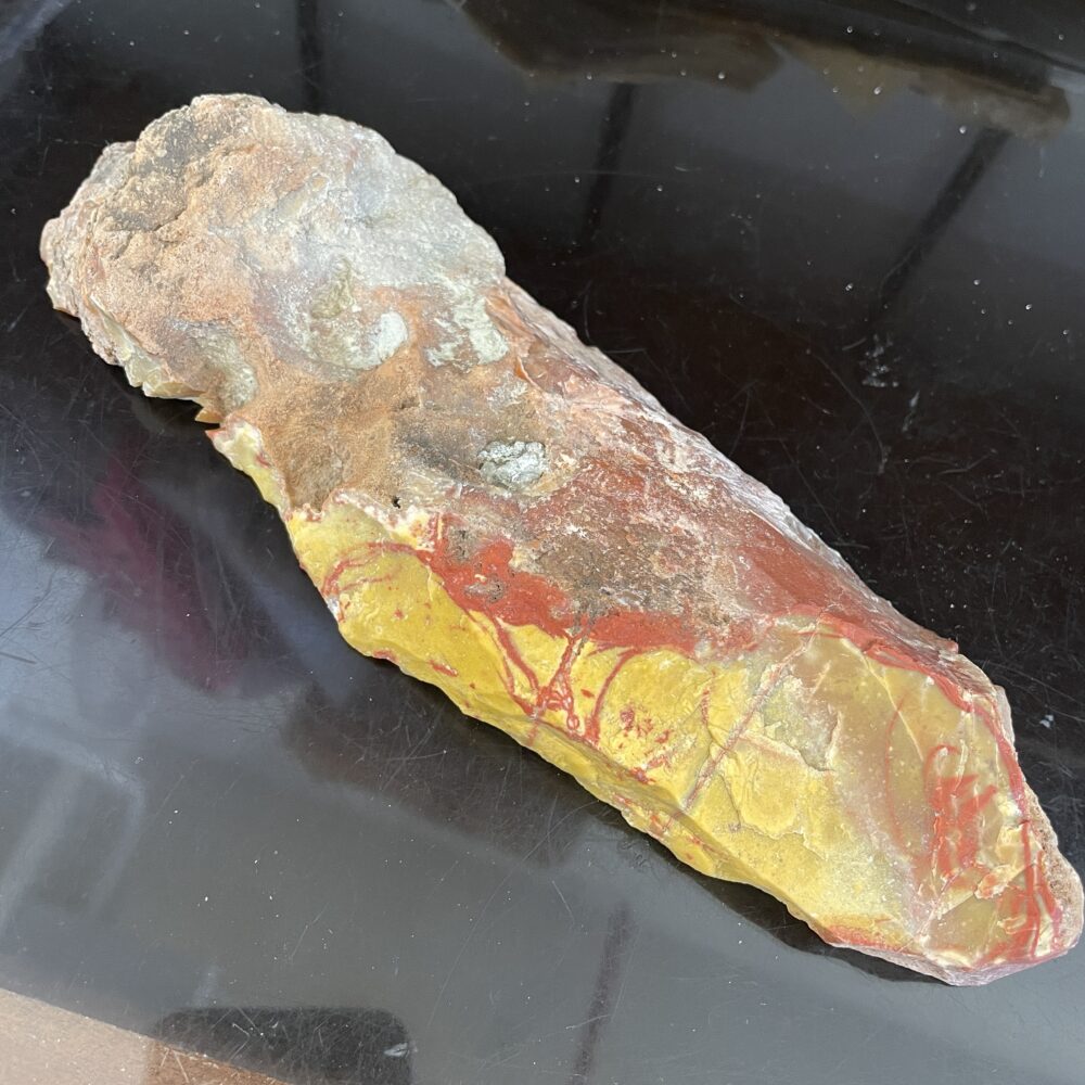 A rough, unpolished stone tool with a yellow, red, and brown surface, displayed on a dark reflective background.