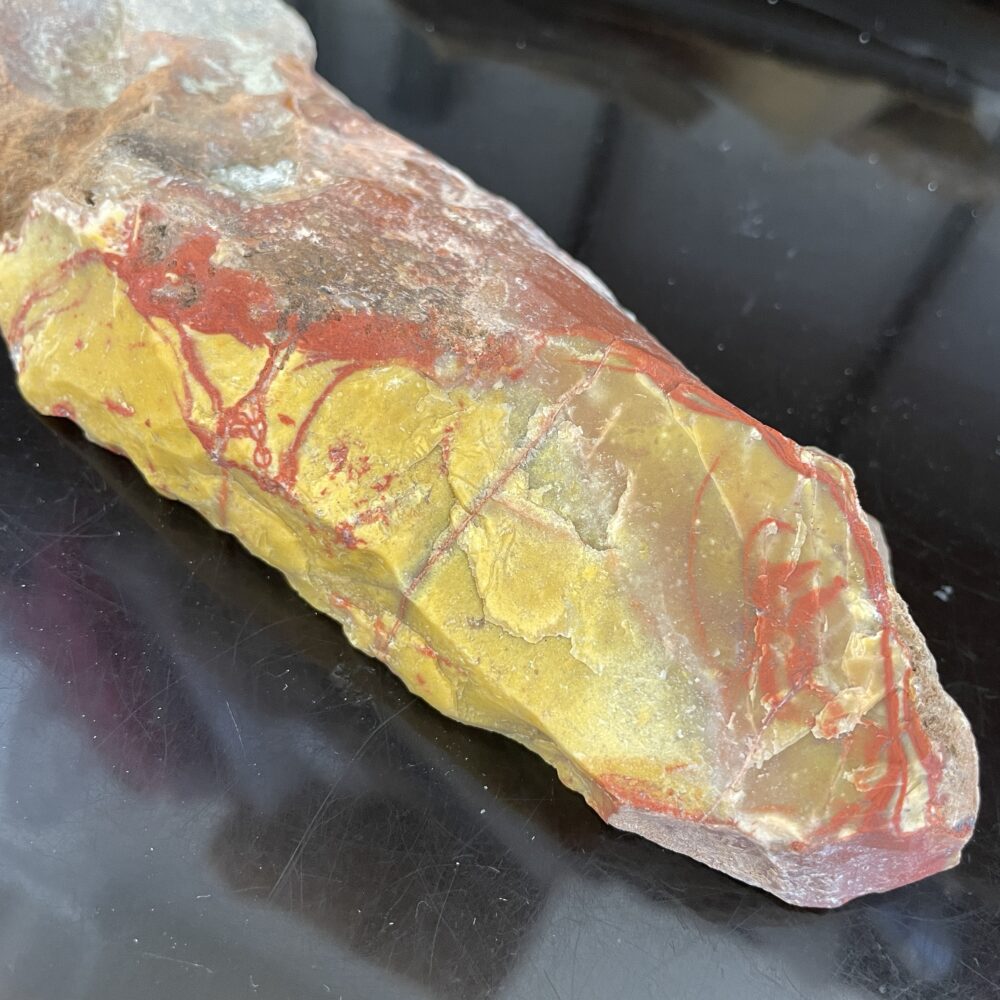 A rough, unpolished piece of jasper with yellow, red, and brown colors sits on a black surface.