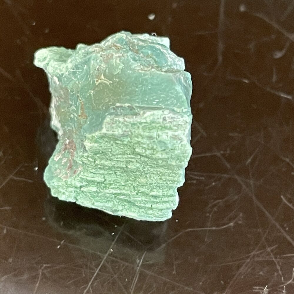 A Rare Green Chromium Petrified Wood specimen with a rough, layered green texture sits on a dark reflective surface.