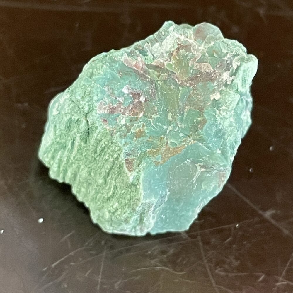 A Rare Green Chromium Petrified Wood specimen featuring an irregular texture with reddish and white patches, displayed on a dark reflective surface.