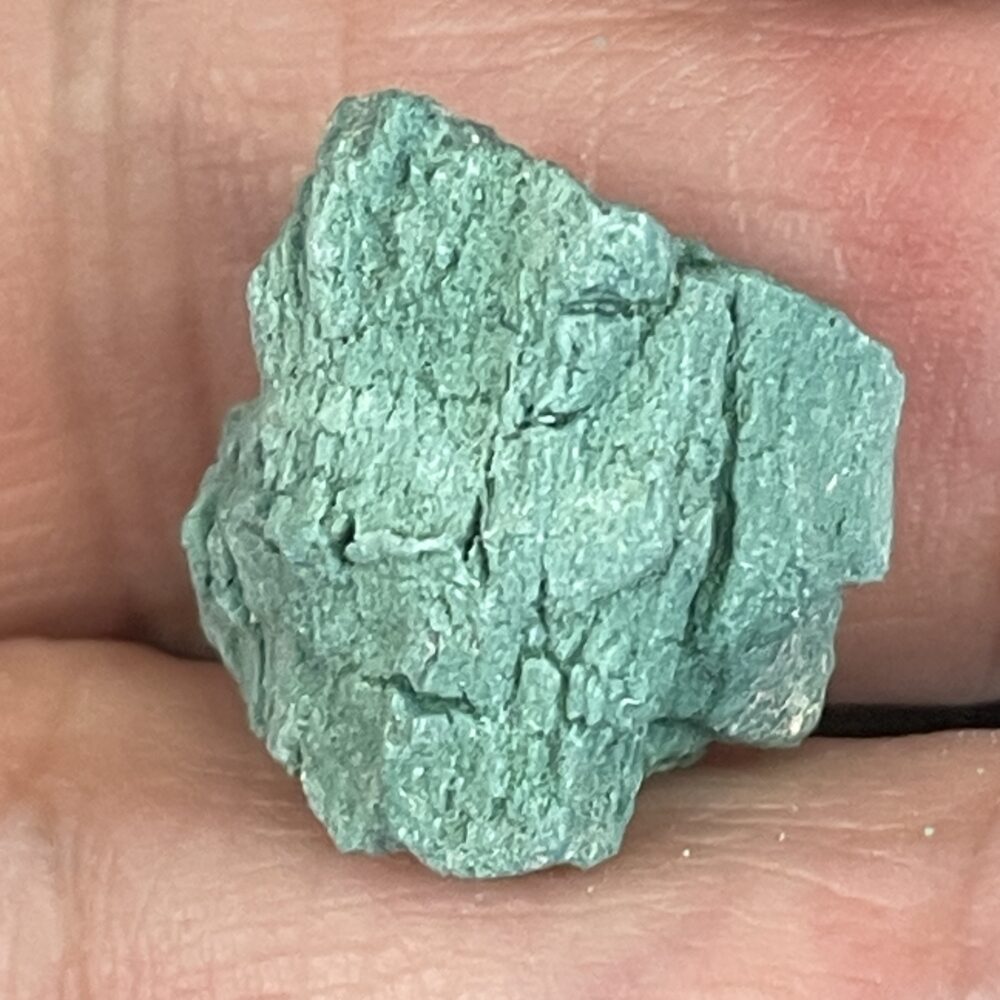 A close-up of Rare Green Chromium Petrified Wood 4.5g, its light green, rough surface delicately held between two fingers.