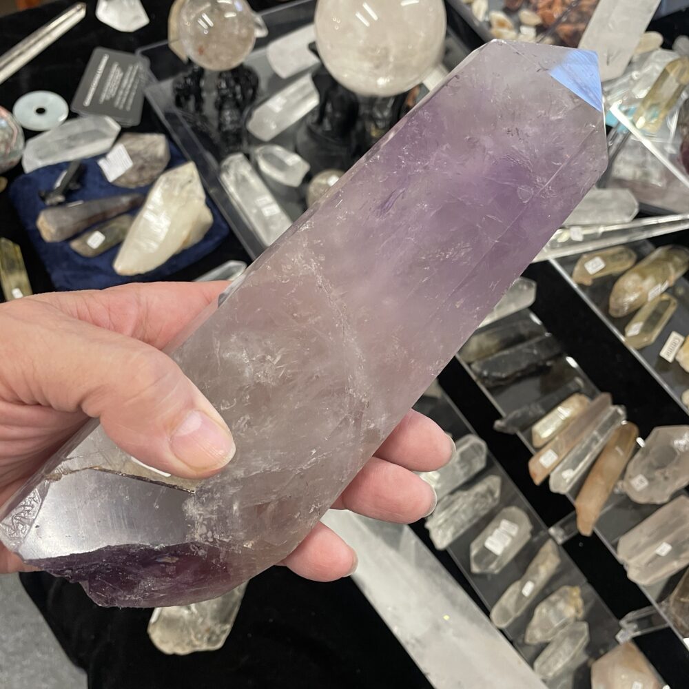 A hand holds a large amethyst crystal point over a display table, showcasing impressive specimens like the Rare 15.4 Inches Smoky Quartz Crystal (5 lbs 10 oz, 2576g) among various other crystals and minerals.