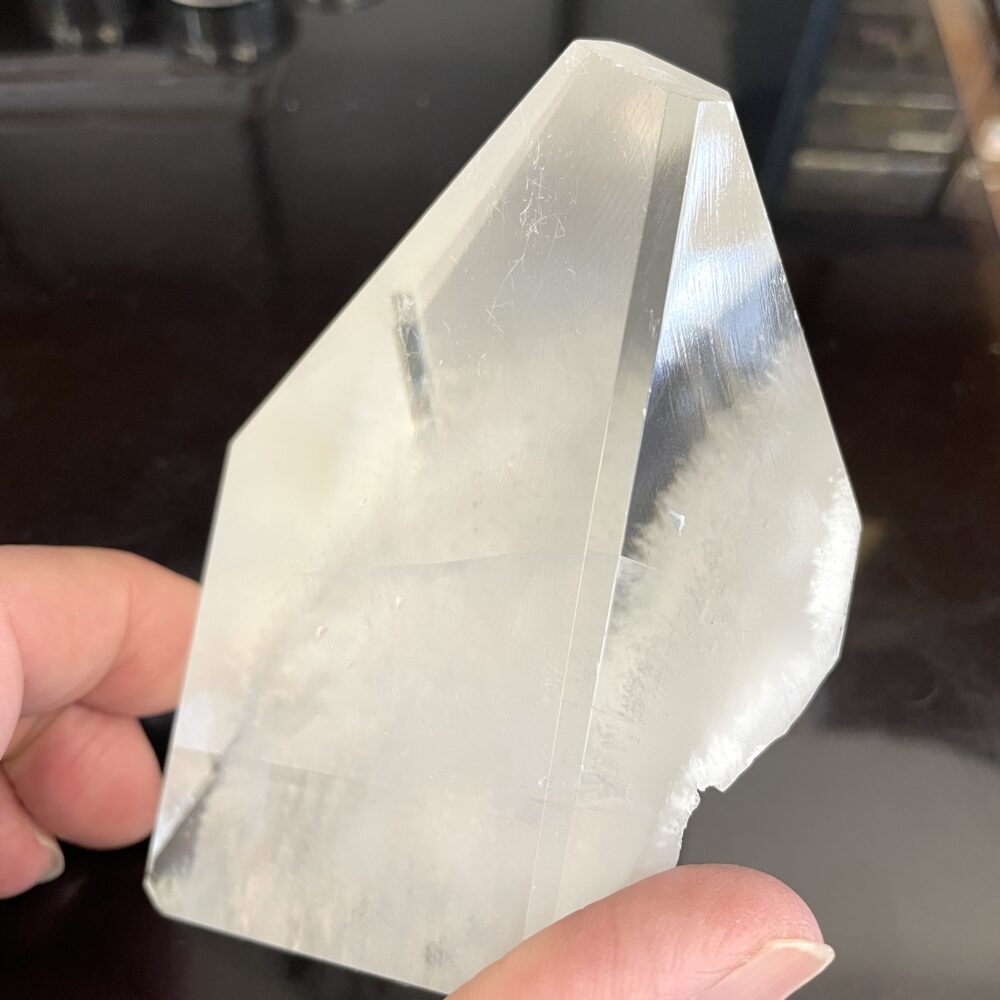 A hand holds a Rare Okenite inside Optical Calcite (Icelandic Spar) crystal from India, 14.7 oz, showing pointed tip and natural facets against a dark background.