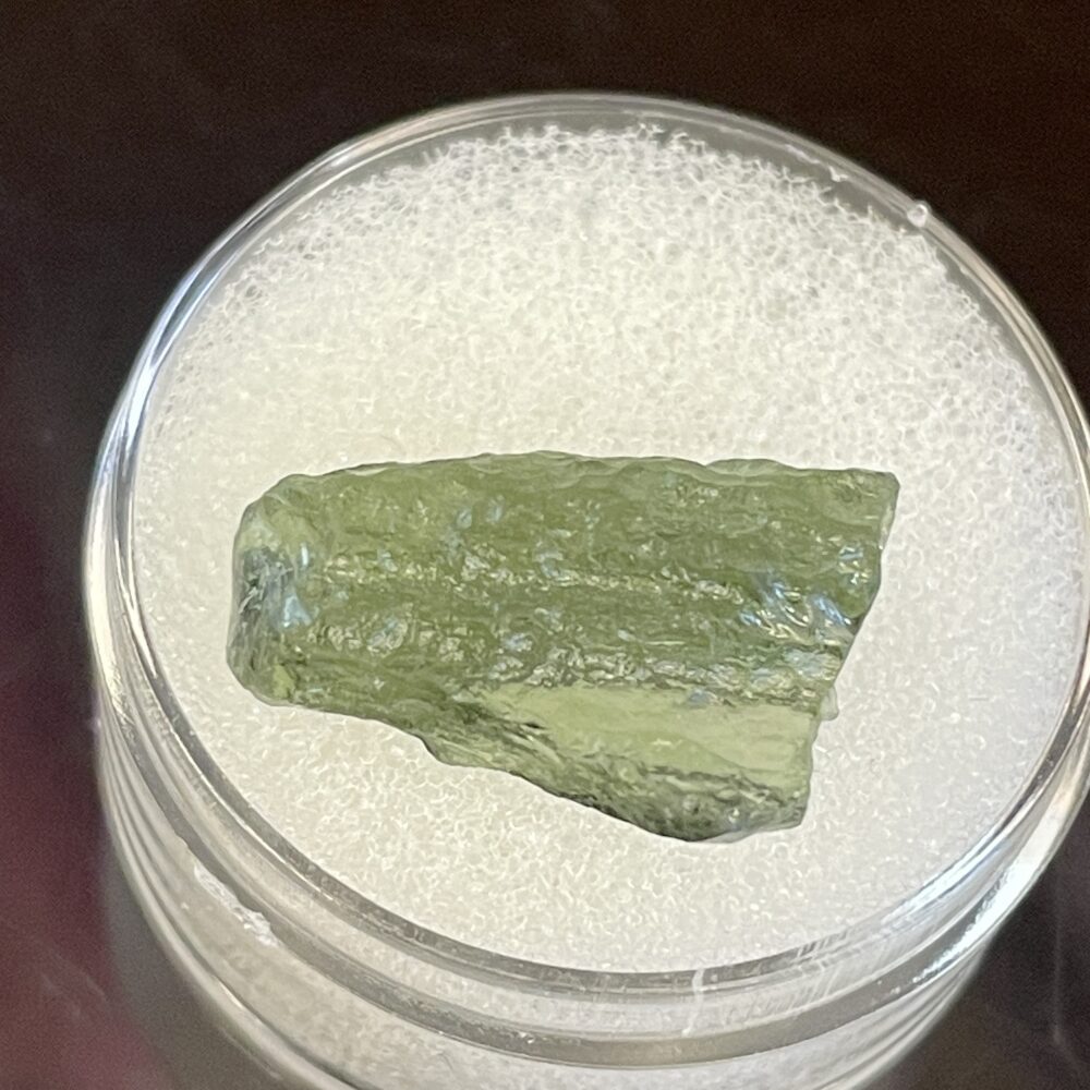 A 0.99 g Moldavite from Chlum, CZ, featuring a green, rough texture, displayed on a white foam pad in a clear plastic container.