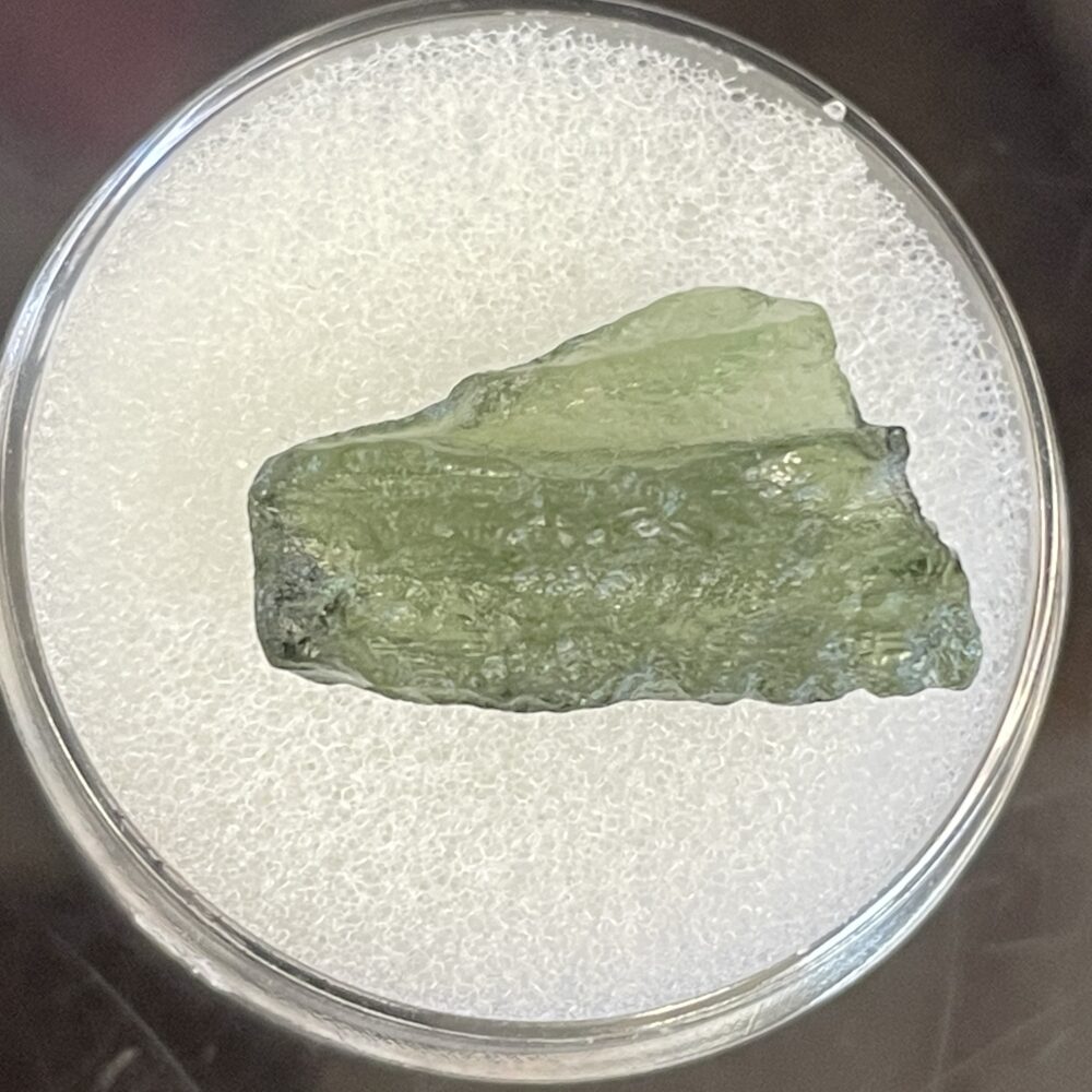 A rough, translucent green Moldavite from Chlum, CZ (0.99g/4.9ct) displayed in a round clear container on a white textured surface.