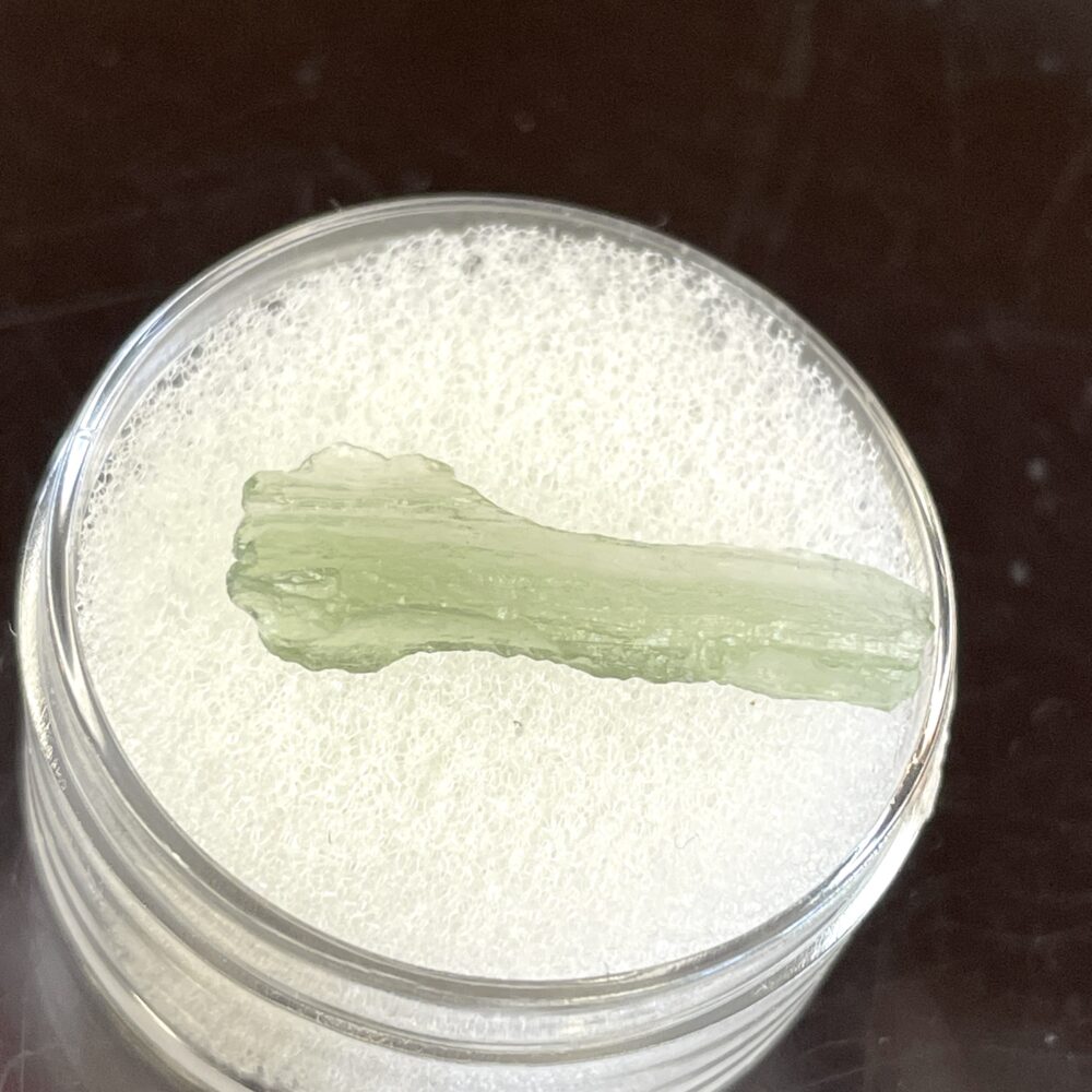 A Moldavite from Chlum in CZ 1.39 g (7.05 ct) rests on a white foam pad inside a clear round container on a dark background.