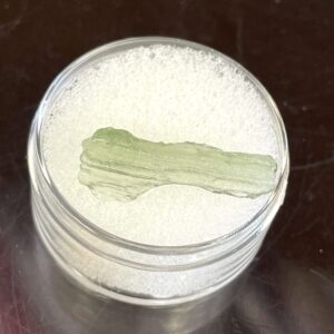 The Moldavite from Chlum in CZ 1.39 g or 7.05 ct rests on white foam inside a clear round container set on a dark surface.