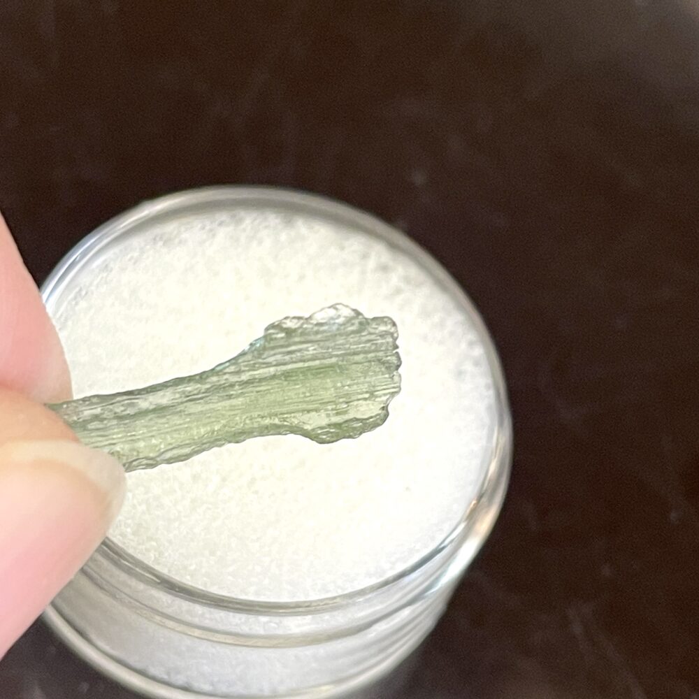 A hand holds a transparent, textured green Moldavite from Chlum in CZ (1.39 g/7.05 ct) above a small, round container of white granules.