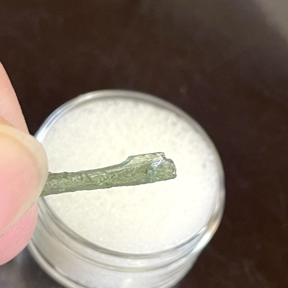 A hand holds a small, green Moldavite from Chlum in CZ (1.39 g / 7.05 ct) above a round container with white cushioning material.