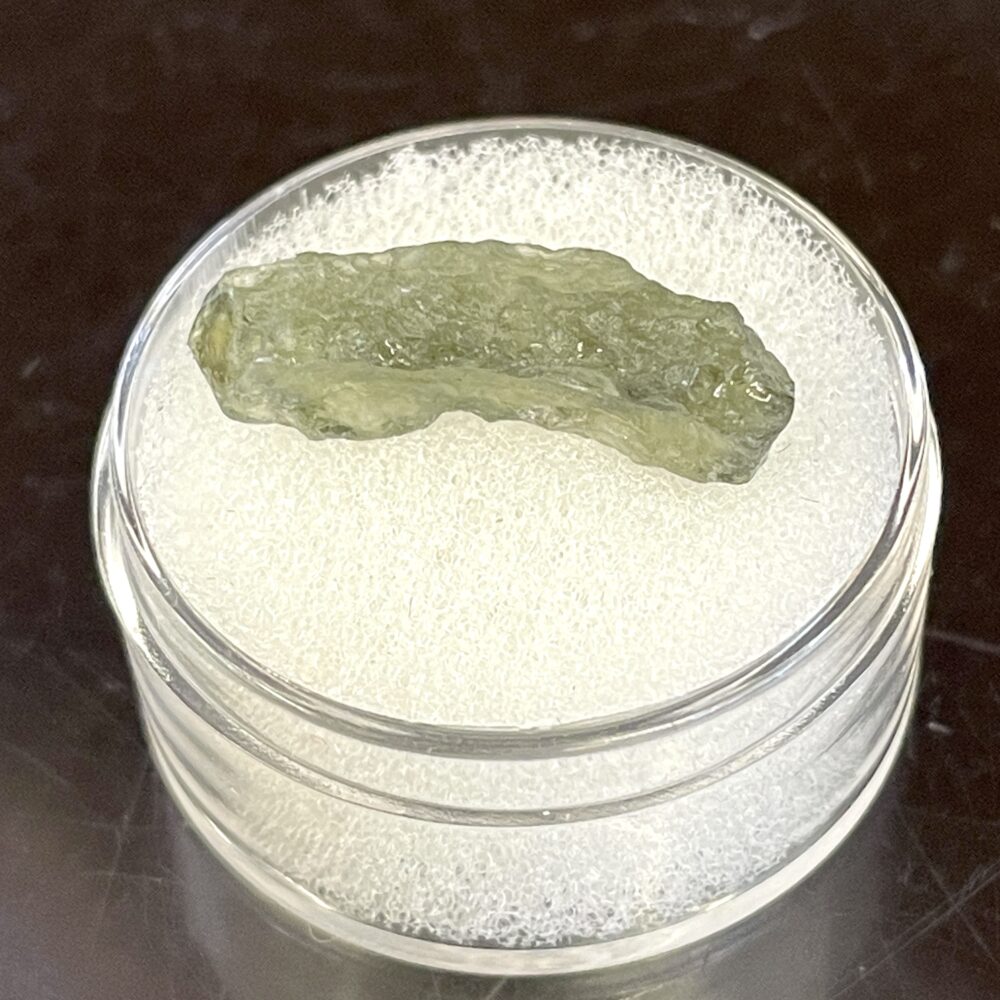 A translucent green Moldavite from Chlum, CZ (0.34g/1.70ct) is displayed on white foam inside a clear round container.