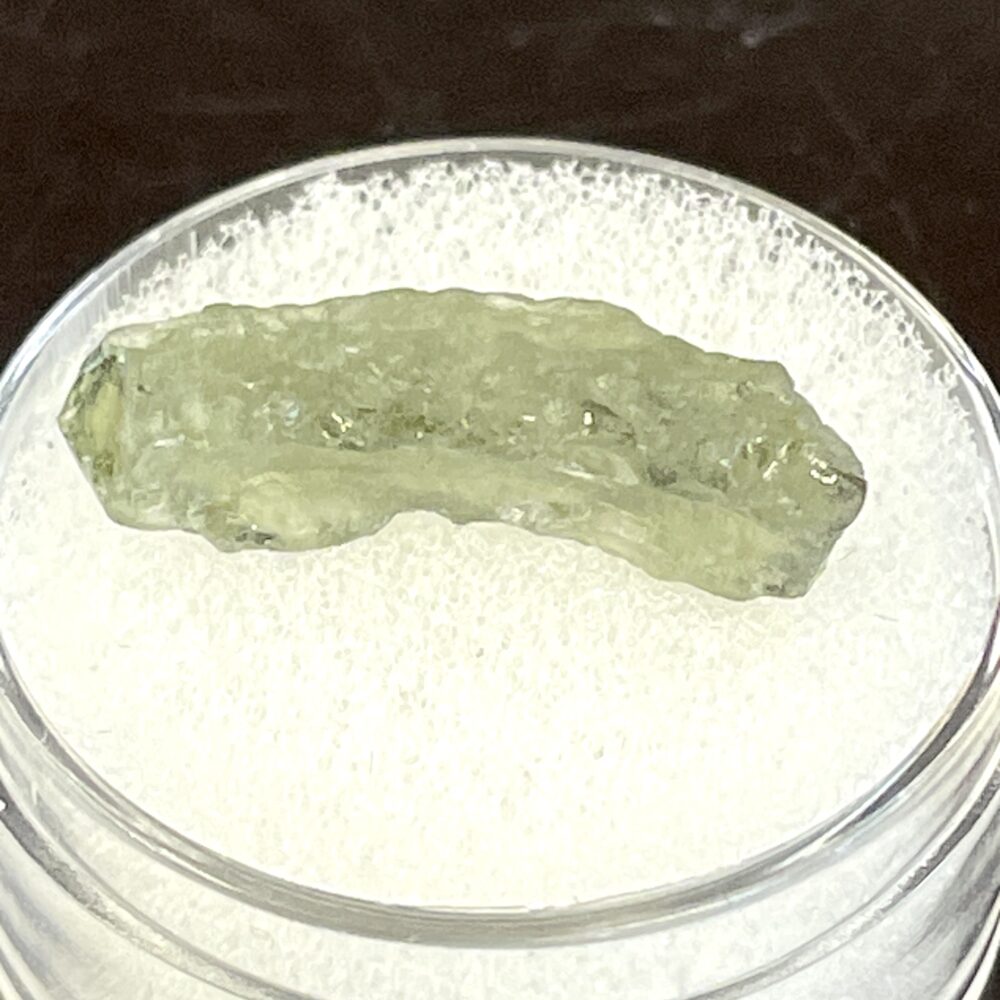 A .34 g (1.70 ct) Moldavite from Chlum, CZ is displayed on white cushioning in a clear plastic container.