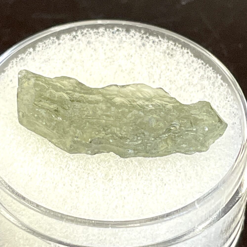 A rough, translucent green Moldavite from Chlum, CZ (.34 g / 1.70 ct) displayed on a white foam pad in a clear circular container.