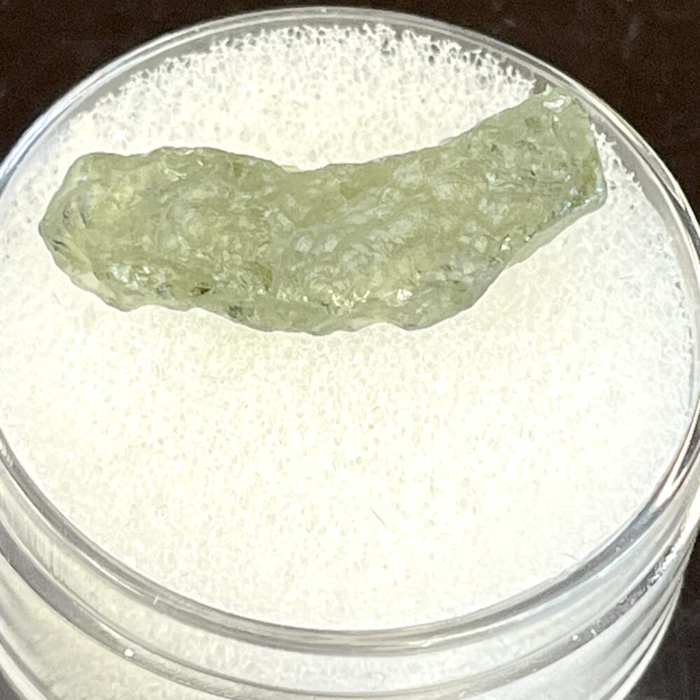 The Moldavite from Chlum in CZ, weighing .34 g / 1.70 ct, is a translucent green mineral on a white foam pad in a clear container.