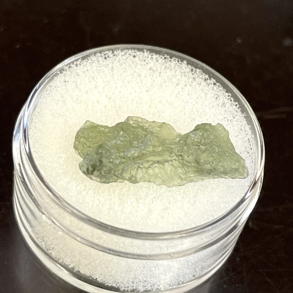A 1.02 g (5.10 ct) Moldavite from Chlum, displayed in a round, clear container lined with white foam.