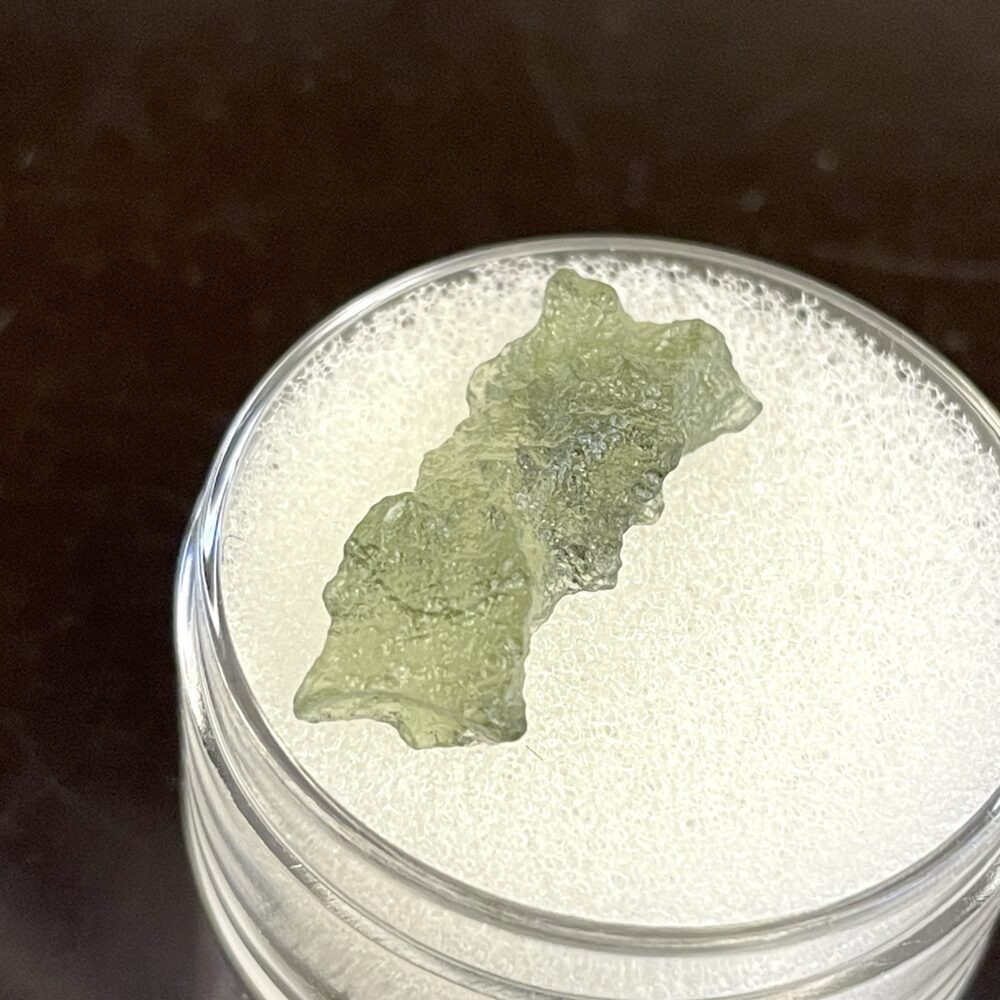 The Moldavite from Chlum in CZ 1.02 g or 5.10 ct is a green, roughly textured specimen on a white foam pad in a clear container.