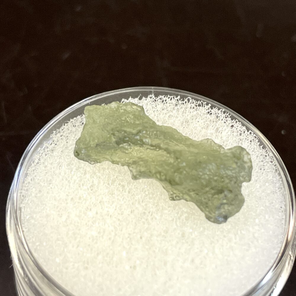 A green Moldavite from Chlum, CZ (1.02 g / 5.10 ct) with an irregular shape sits on a white foam pad in a clear round container.