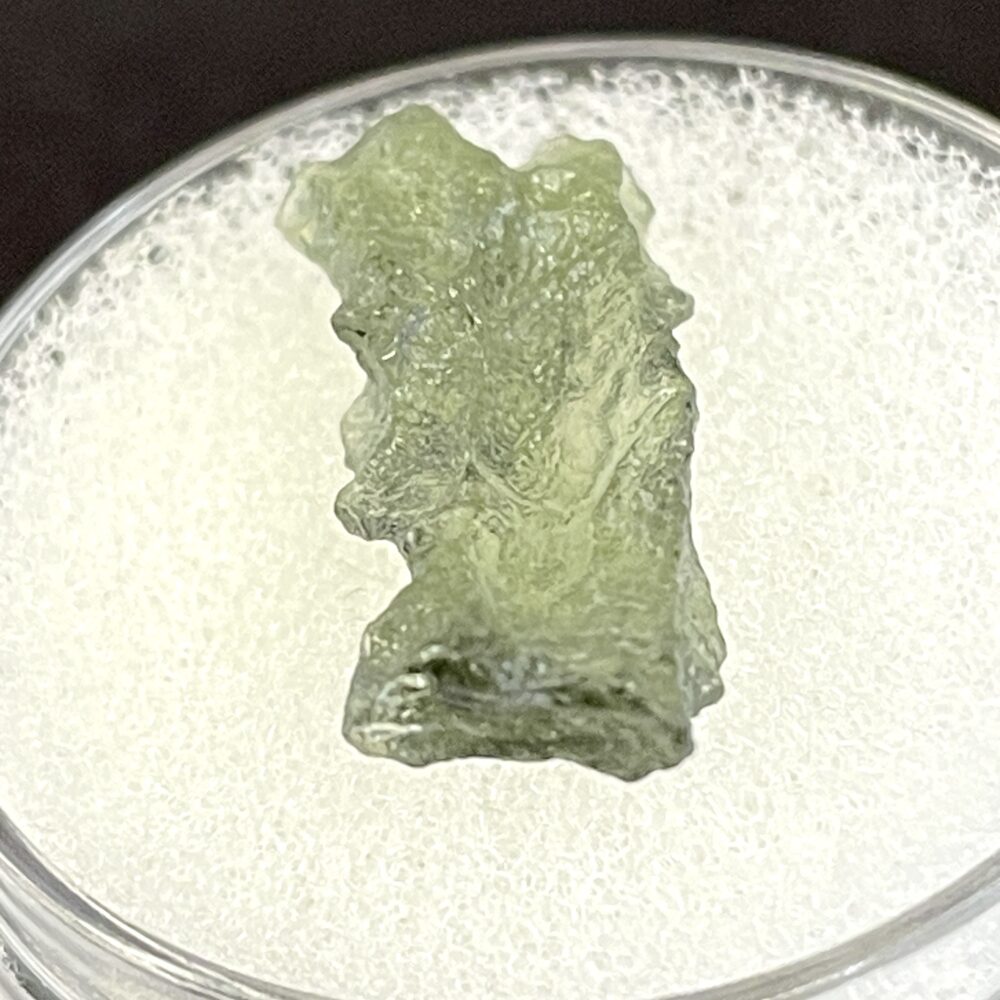 A rough, translucent Moldavite from Chlum, CZ (1.02 g/5.10 ct) is displayed on a textured white surface inside a round container.