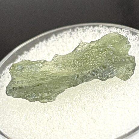 A translucent, rough Moldavite from Chlum, CZ (1.02 g/5.10 ct) is displayed on a white textured surface in a clear container.