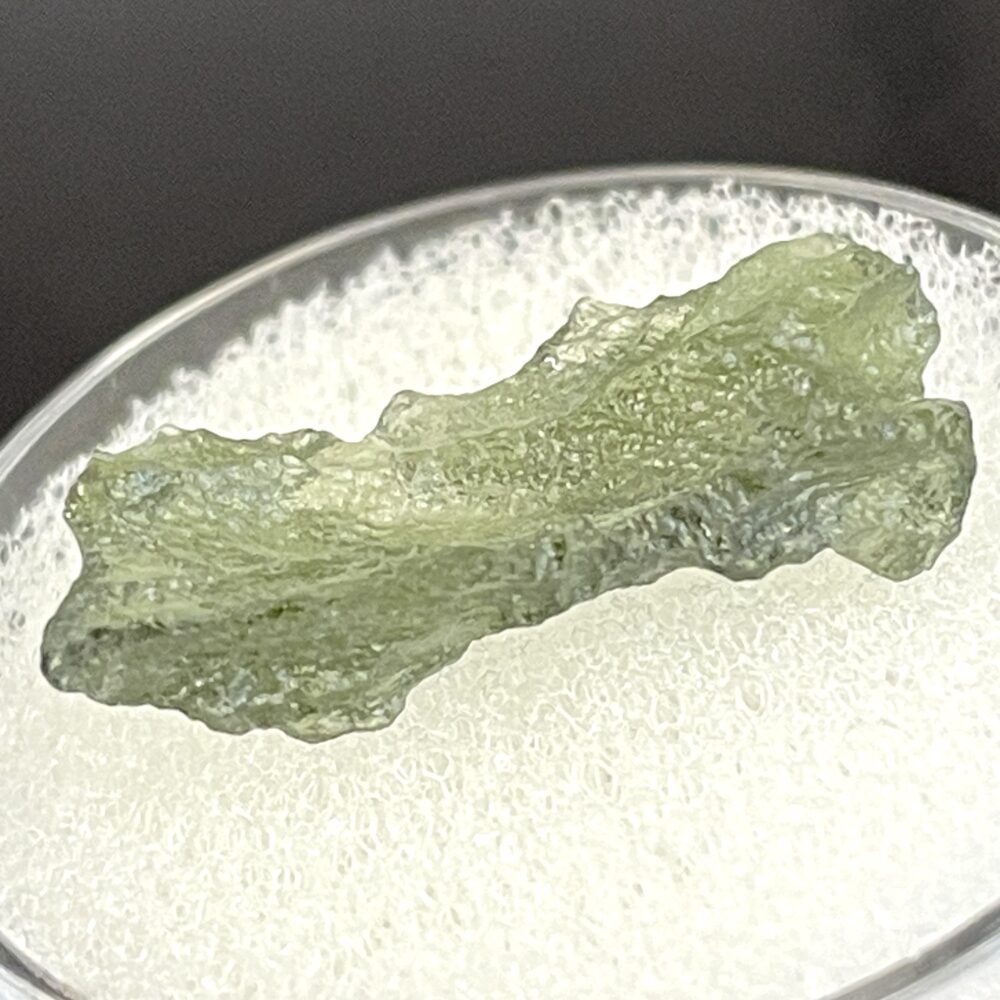 A translucent, rough Moldavite from Chlum, CZ (1.02 g/5.10 ct) is displayed on a white textured surface in a clear container.