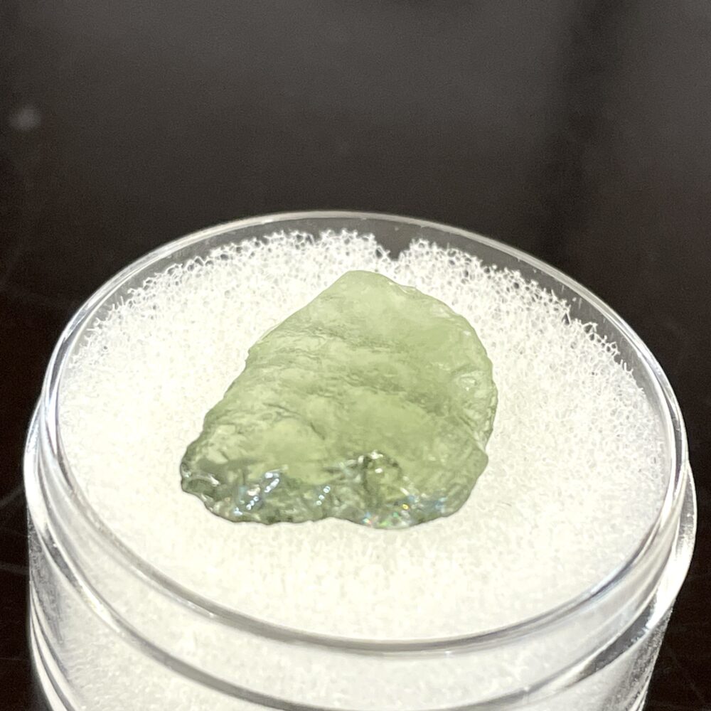 A translucent green Moldavite from Chlum, CZ (1.02 g / 5.10 ct) is displayed on white foam in a clear round container.