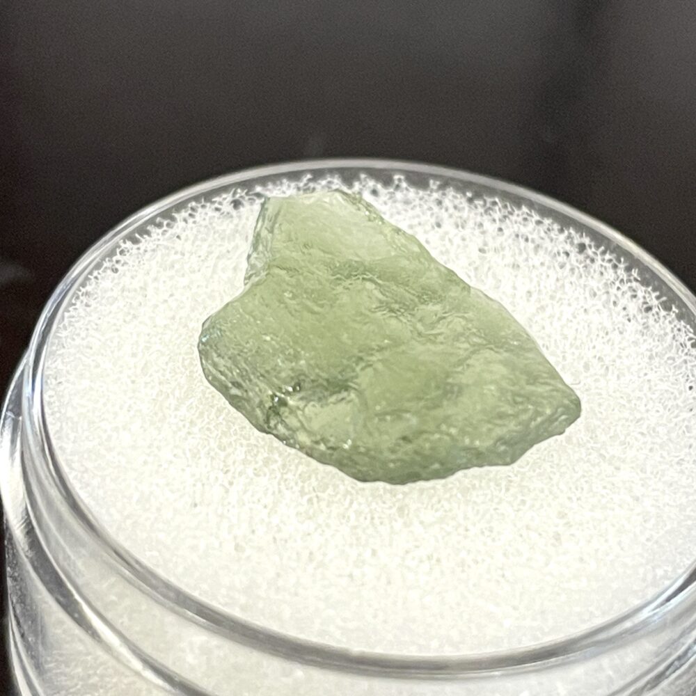A translucent green Moldavite from Chlum, CZ (1.02 g / 5.10 ct) is showcased on a white textured surface in a clear container.