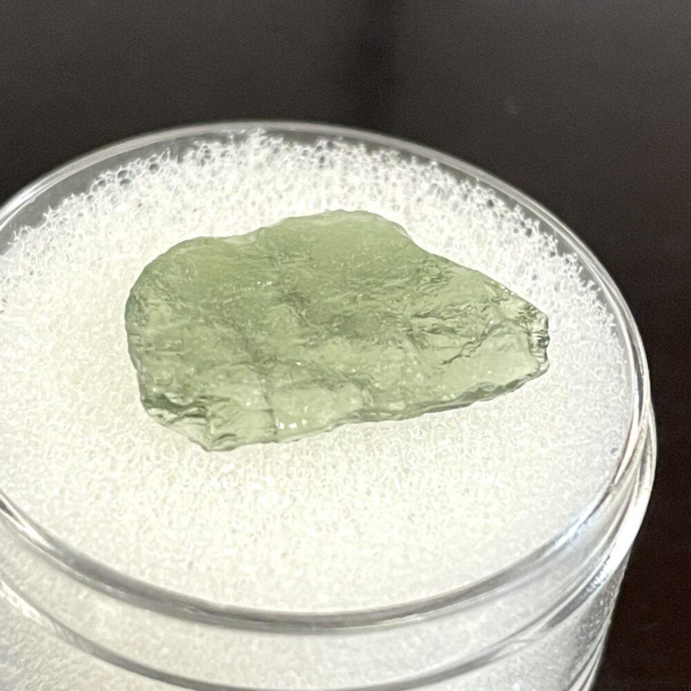 A translucent green Moldavite from Chlum in CZ, 1.02 g, rests on a white foam pad inside a clear round container.