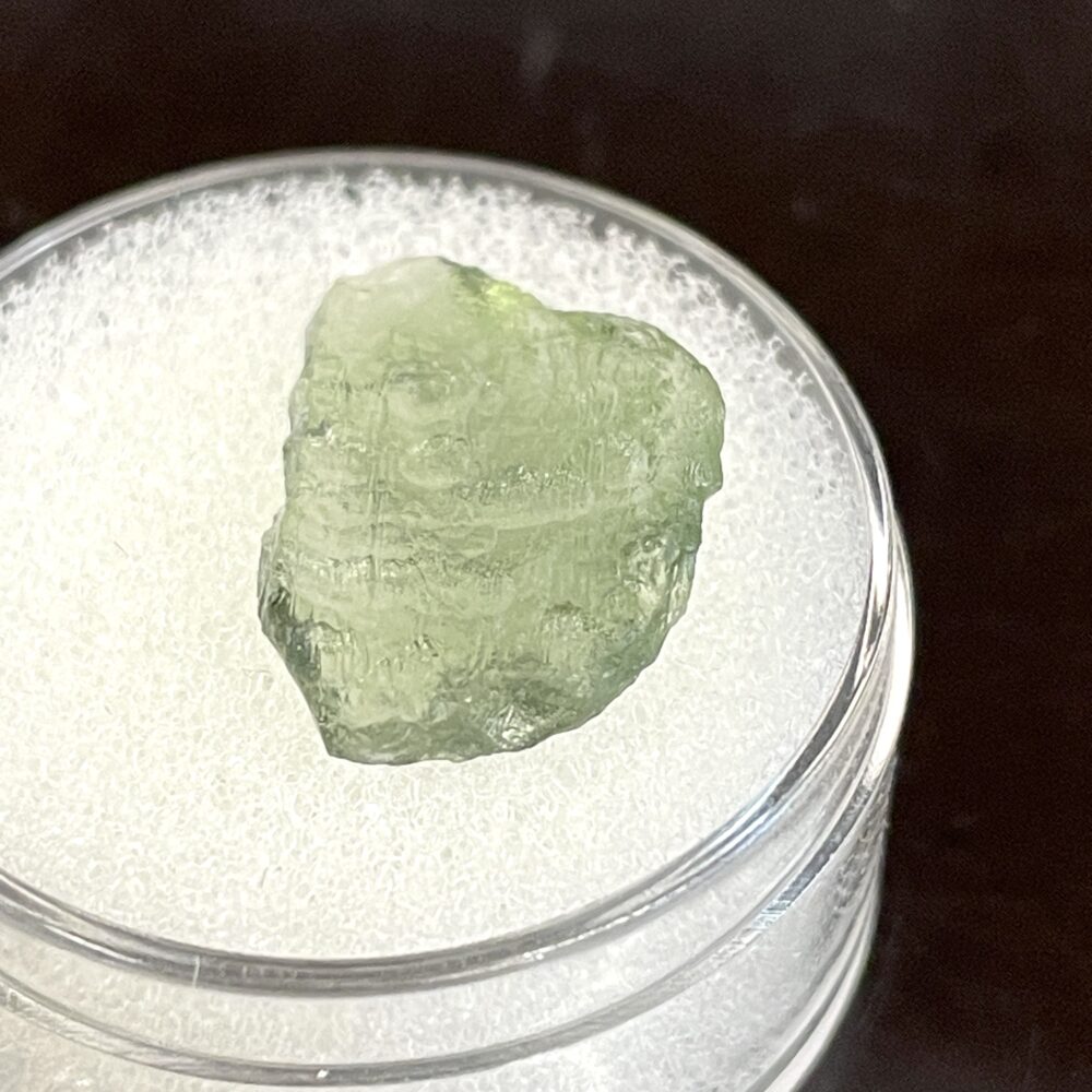A rough, translucent green Moldavite from Chlum, CZ (1.02 g / 5.10 ct), displayed on a white foam pad in a clear round container.