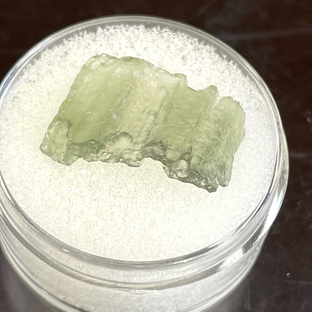 A rough, green Moldavite from Chlum (0.69 g, 3.45 ct) is displayed upright in a round clear container with white foam padding.