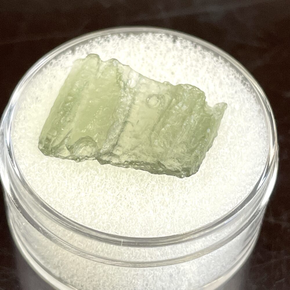 A rough, light green Moldavite from Chlum, CZ (.69 g/3.45 ct), displayed on white foam in a clear round container.