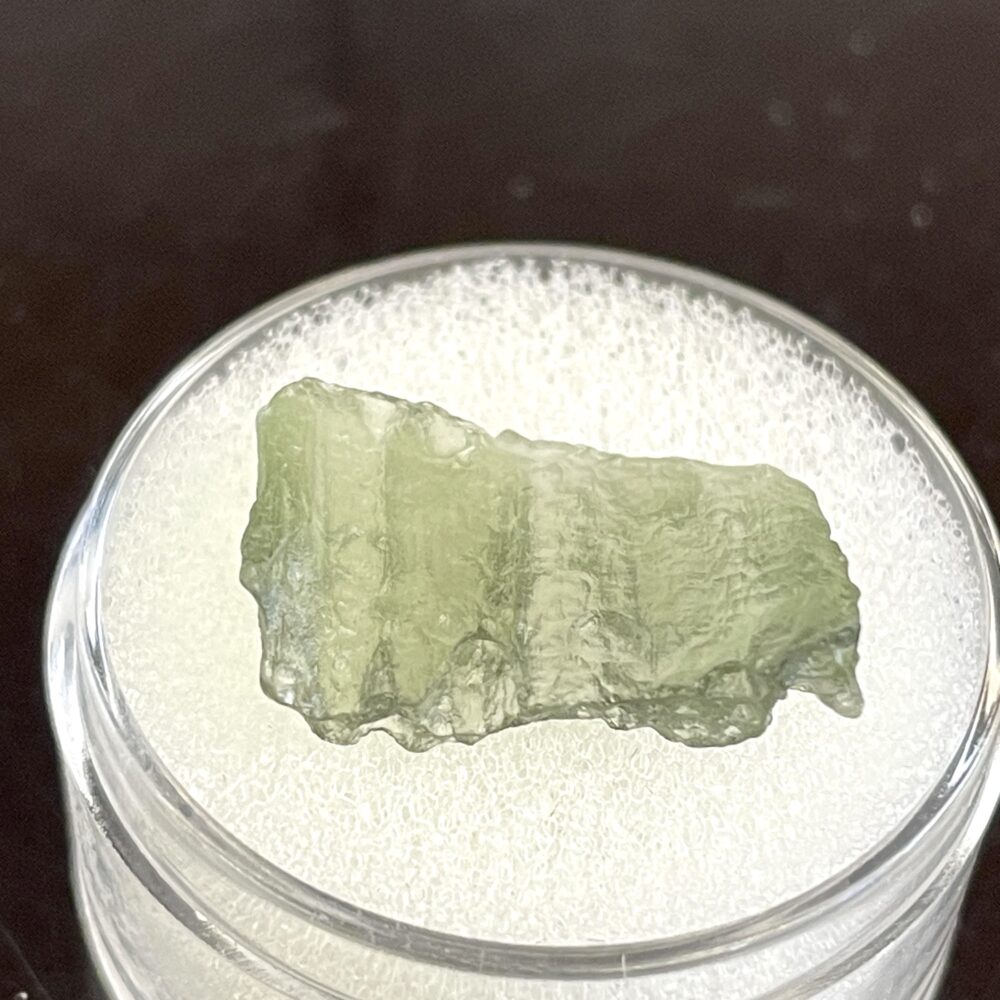 A .69 g (3.45 ct) Moldavite from Chlum, CZ, displayed in a clear round container with white padding.