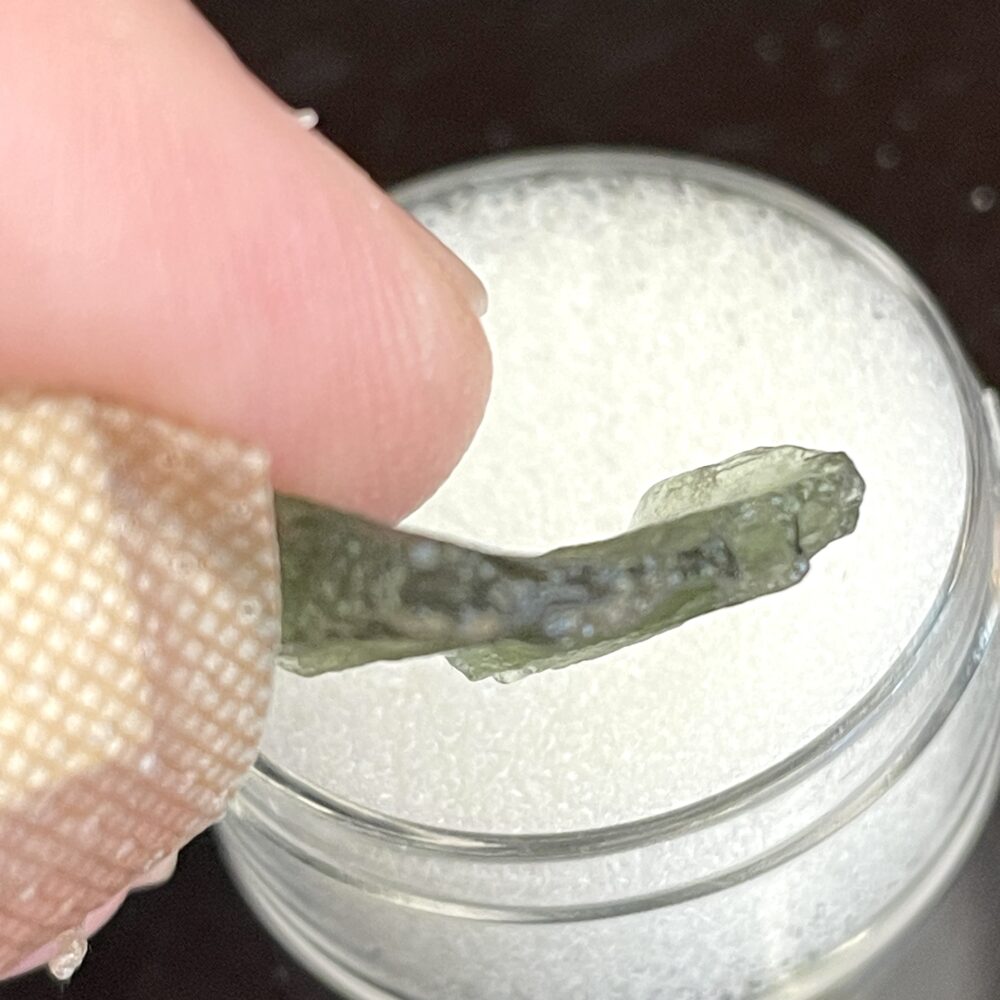 A close-up shows a hand holding a .69 g (3.45 ct) Moldavite from Chlum, CZ, above a white circular container.