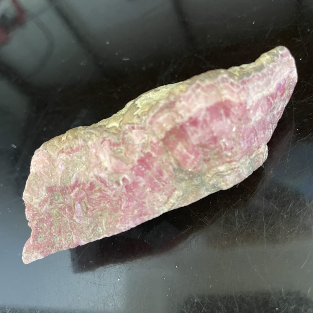 A beautiful old stock Rhodochrosite specimen with natural, uneven edges and yellowish tones, displayed on a black surface.