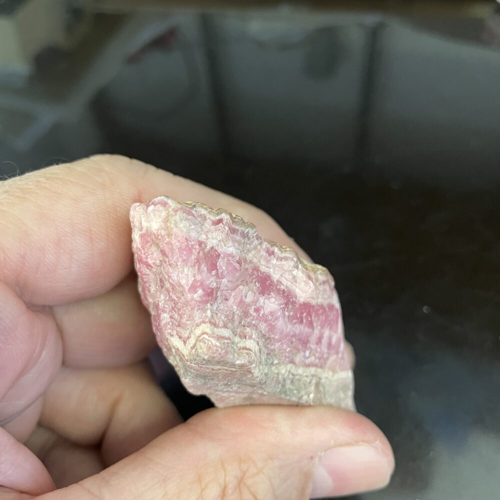 A hand holds a Rhodochrosite old stock specimen featuring a rough, pink and white jagged surface against a dark background.