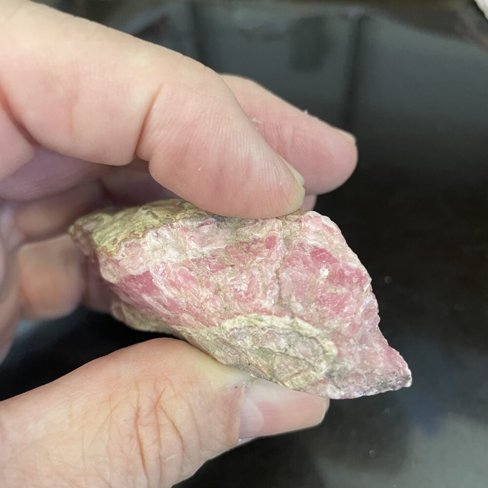 A hand holds the Rhodochrosite Beautiful Old Stock Specimen against a dark background—an intriguing addition to any collection.