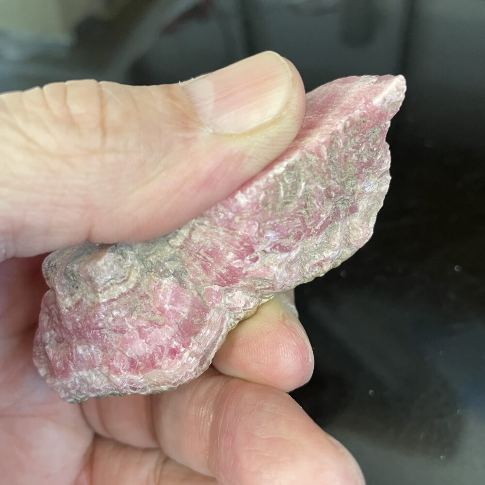 A hand holds the Rhodochrosite Beautiful Old Stock Specimen with grayish-white patches, displayed against a dark background.