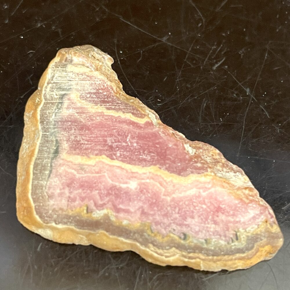 A polished slice of pink and brown banded Rhodochrosite old stock specimen is displayed on a dark reflective surface.