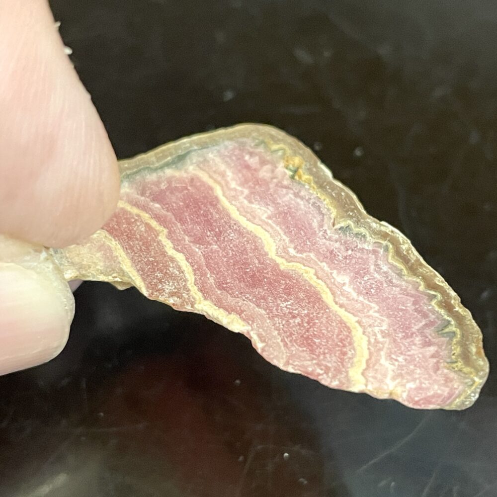 A hand holds a thin, translucent slice of pink and white banded Rhodochrosite specimen against a dark background.