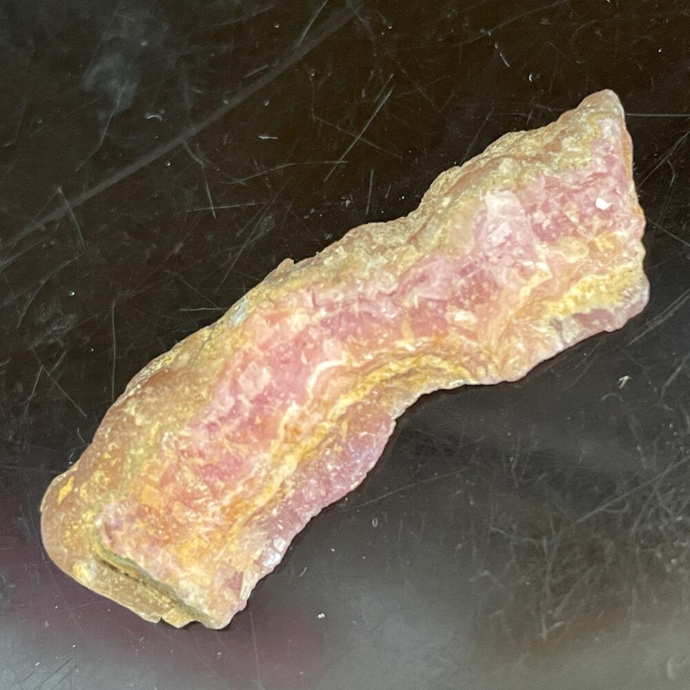A rough, unpolished Rhodochrosite specimen with pink and yellow hues rests on a dark, reflective surface, highlighting its charm.
