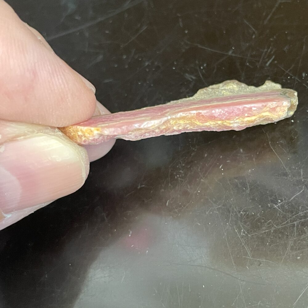 A hand holds a thin, irregular slice of Rhodochrosite Beautiful Old Stock Specimen 6.71 grams against a dark background.