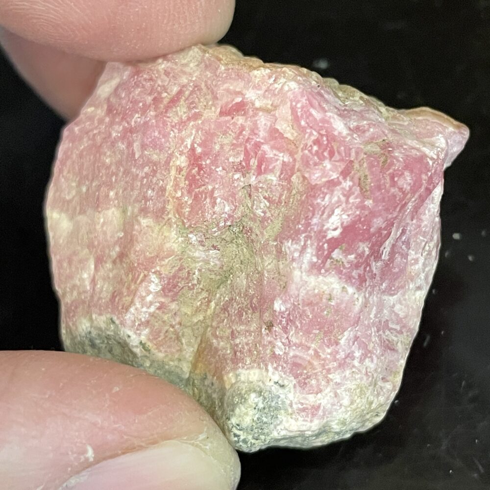 A close-up of fingers holding a 7.38 gram Rhodochrosite specimen with a rough pink surface and white-green areas on black.