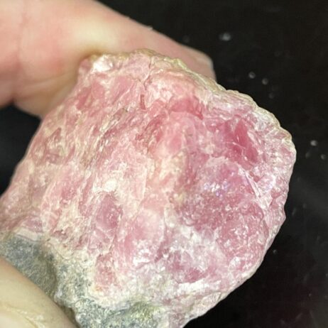 A close-up of a hand holding the Rhodochrosite Beautiful Old Stock Specimen 7.38 grams, highlighting its rough, unpolished textures against a black background.