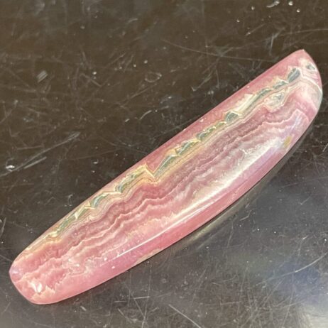 A polished, elongated Rhodochrosite stone with pink and white wavy bands is displayed on a dark surface—7.38 grams.
