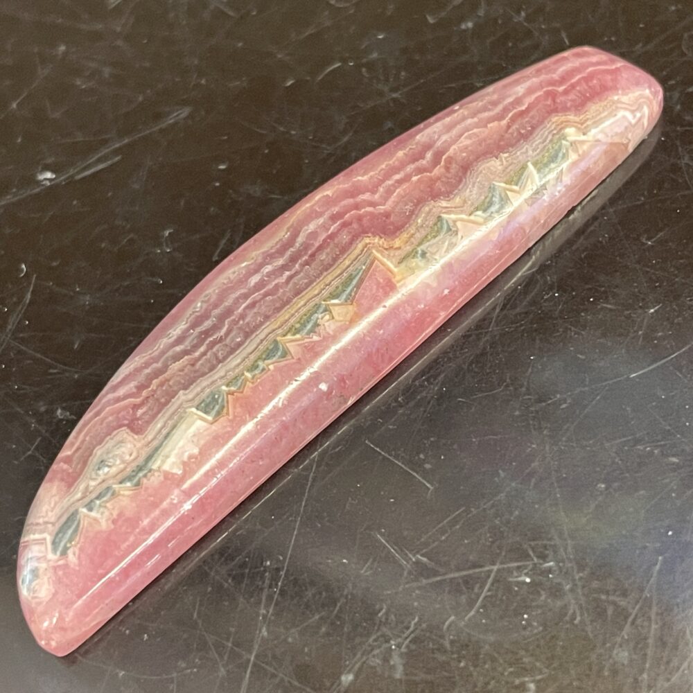 A beautiful 7.38-gram Rhodochrosite Old Stock Specimen with pink and cream bands and a curved, elongated shape rests on a dark surface.