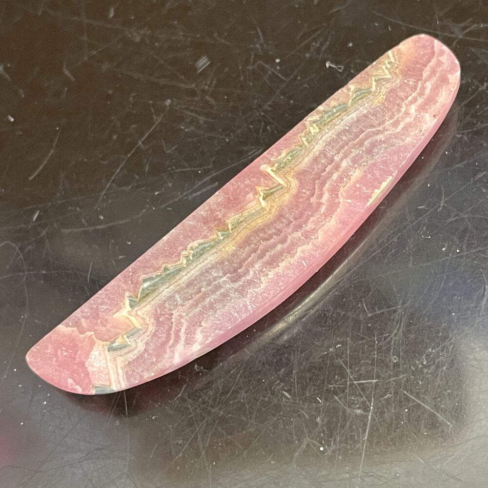 A polished Rhodochrosite old stock specimen, 7.38g, with pink, white, and tan banding on a reflective dark surface.
