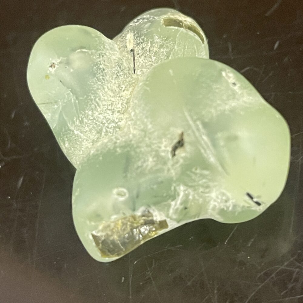 A close-up of a translucent, greenish Prehinite Natural Shaped Tumble Polished crystal showing a glossy surface and internal inclusions.