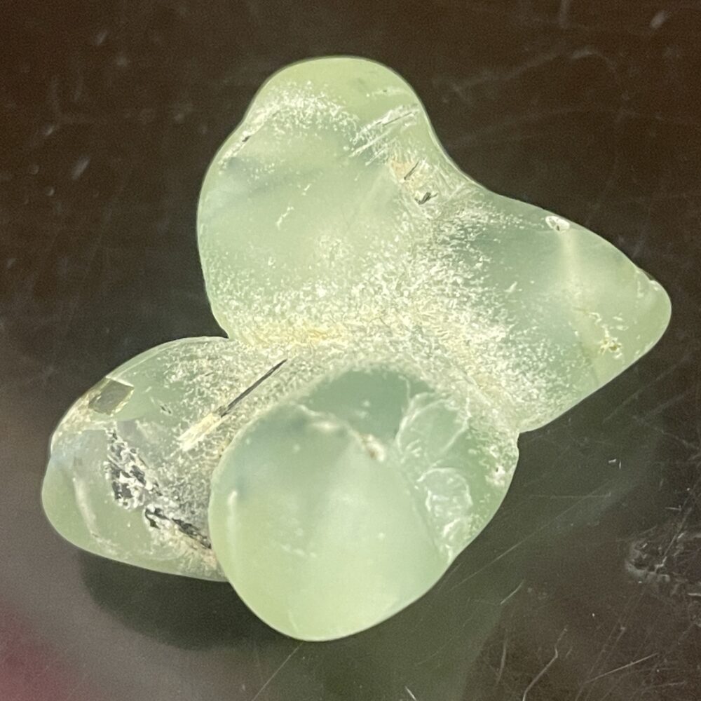A translucent, light green Prehnite tumble-polished stone (21.18g / .747 oz) with a smooth finish rests on a dark, reflective surface.